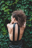 A person with curly brown hair viewed from the back, wearing a black halter top dress. The background consists of dense green foliage.