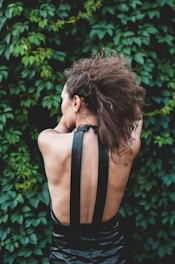 A person with curly brown hair viewed from the back, wearing a black halter top dress. The background consists of dense green foliage.