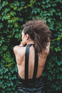 A person with curly brown hair viewed from the back, wearing a black halter top dress. The background consists of dense green foliage.