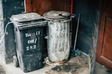 Two trash bins, one black plastic with a rectangular lid and the other a metallic barrel, are placed next to each other against a dark wall. A red wooden door and a broom are visible nearby, with scattered debris and dirt on the ground.