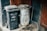 Two trash bins, one black plastic with a rectangular lid and the other a metallic barrel, are placed next to each other against a dark wall. A red wooden door and a broom are visible nearby, with scattered debris and dirt on the ground.