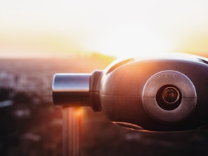 A close-up view of a telescope against a blurred cityscape background with a bright sunset, creating a warm, glowing effect.