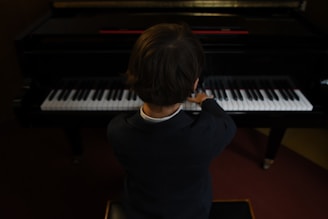 A child in formal attire is seated at a piano, playing the keys with both hands. The perspective is from behind, focusing on the posture and concentration of the child as they engage with the musical instrument. The piano is in a dimly lit room, creating an intimate atmosphere.