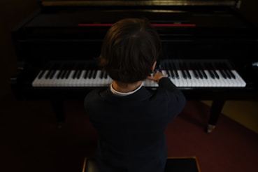 A child in formal attire is seated at a piano, playing the keys with both hands. The perspective is from behind, focusing on the posture and concentration of the child as they engage with the musical instrument. The piano is in a dimly lit room, creating an intimate atmosphere.