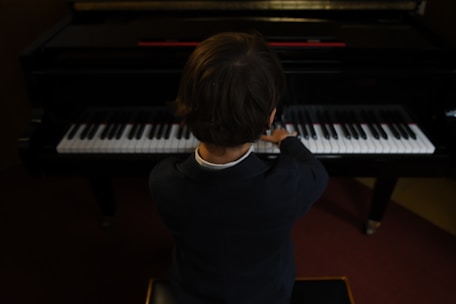 A child in formal attire is seated at a piano, playing the keys with both hands. The perspective is from behind, focusing on the posture and concentration of the child as they engage with the musical instrument. The piano is in a dimly lit room, creating an intimate atmosphere.