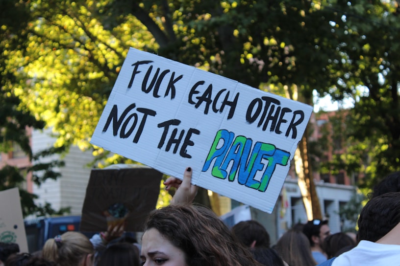 People gathered in a public space holding a protest sign with a message about environmental concerns. Trees and buildings are visible in the background, indicating an outdoor setting. The focus is on a sign with bold black letters and colorful green and blue accents.
