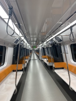 An empty train car with orange and white seating, illuminated by overhead lighting. The view is down the center aisle with metal poles for standing passengers. The interior has a modern and clean design, with a digital display indicating a station name.