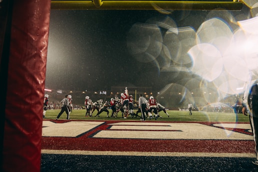 A rainy football game viewed from the end zone, with players lined up on the field. Bright stadium lights illuminate the scene and water droplets create light refractions in the foreground.