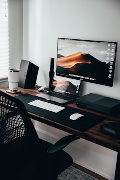 A minimalist desk setup featuring a large monitor displaying a desert landscape, a closed laptop with a keyboard and mouse on a black desk mat, a white tumbler, a black office chair, a desktop PC, and a set of cables.