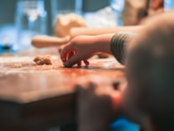 A child's hand reaching for small pieces of dough on a wooden table with a blurred background. The scene suggests a playful and creative activity, possibly related to baking or crafting.