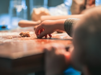 A child's hand reaching for small pieces of dough on a wooden table with a blurred background. The scene suggests a playful and creative activity, possibly related to baking or crafting.