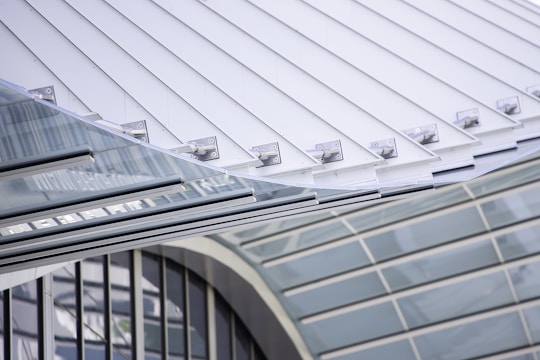 A modern architectural structure featuring sleek lines and a combination of metal and glass elements. The design incorporates a series of metal panels and glass surfaces with visible bolts and support elements, showcasing an industrial yet sophisticated aesthetic.