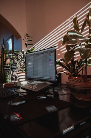 A workspace is illuminated by natural light streaming through blinds, casting striped shadows on the wall. A computer monitor displays an application, surrounded by lush potted plants. The atmosphere is cozy and warm, with a mix of technology and nature.