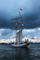 A large sailing ship with multiple masts is navigating through a body of water. The sky is overcast with dark clouds, adding a dramatic backdrop. Flags are flying at the top of the masts, and several people are visible on the deck near the stern. The water appears slightly choppy, reflecting the moody atmosphere.