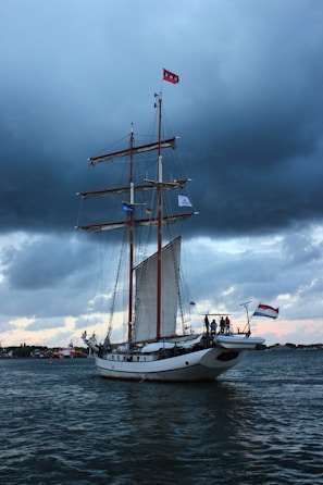 A large sailing ship with multiple masts is navigating through a body of water. The sky is overcast with dark clouds, adding a dramatic backdrop. Flags are flying at the top of the masts, and several people are visible on the deck near the stern. The water appears slightly choppy, reflecting the moody atmosphere.