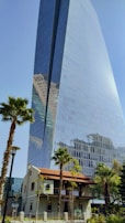 A tall, modern skyscraper with a glass facade reflecting nearby buildings and the sky. Surrounding the base of the building, there are several palm trees and a two-story structure with a red-tiled roof. The scene combines urban architecture with natural elements.