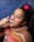 A person wearing makeup with blue eyeshadow and bold lipstick is depicted flossing their teeth. They are wearing a colorful striped sweater and have a pink scrunchie in their hair against a textured blue background.