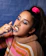 A person wearing makeup with blue eyeshadow and bold lipstick is depicted flossing their teeth. They are wearing a colorful striped sweater and have a pink scrunchie in their hair against a textured blue background.