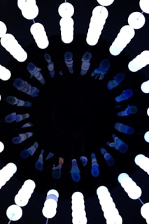 A circular arrangement of vertical fluorescent lights, glowing brightly against a dark background. The lights are tube-shaped and emit a bluish-white hue, creating a contrast with the surrounding darkness.