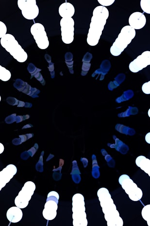 A circular arrangement of vertical fluorescent lights, glowing brightly against a dark background. The lights are tube-shaped and emit a bluish-white hue, creating a contrast with the surrounding darkness.