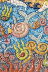 Colorful mosaic art depicts swirling patterns resembling underwater sea life with abstract shapes. The artwork features vibrant tiles in blue, yellow, green, and orange, with designs that mimic seashells, seaweed, and waves.