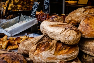 A variety of baked goods are displayed, including loaves of artisanal bread, croissants, and cinnamon rolls with cream cheese frosting. The scene is bustling, likely at a bakery or market stall.