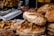 A variety of baked goods are displayed, including loaves of artisanal bread, croissants, and cinnamon rolls with cream cheese frosting. The scene is bustling, likely at a bakery or market stall.