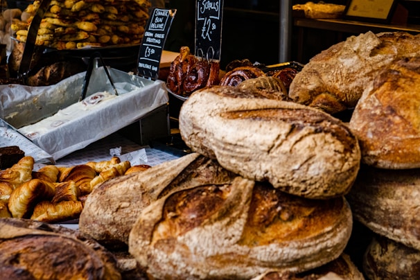 A variety of baked goods are displayed, including loaves of artisanal bread, croissants, and cinnamon rolls with cream cheese frosting. The scene is bustling, likely at a bakery or market stall.