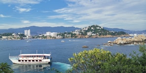 A white tour boat is moving across a blue bay, with several passengers on board. In the background, there is a coastal city with tall buildings, set against a backdrop of green hills and mountains. The sky is partly cloudy, and there are a few small boats in the water. In the foreground, lush green foliage is visible along the water's edge.