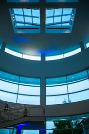 A modern interior space with large, curved glass windows and a ceiling featuring skylights. The structure includes a metal staircase and visible supporting beams, complemented by ambient blue lighting.