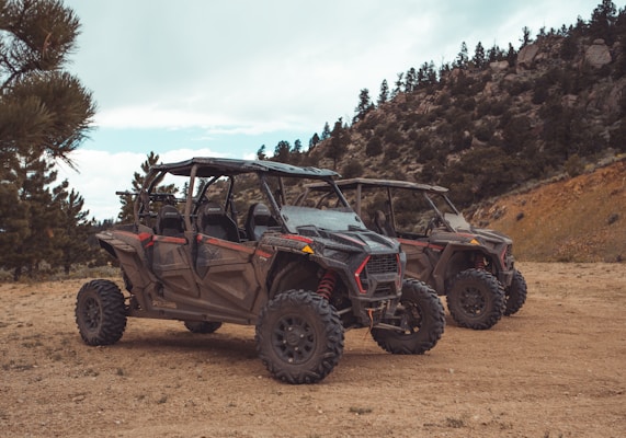 Two off-road vehicles are parked on a dirt path surrounded by a natural landscape featuring pine trees and rocky hills. The vehicles have rugged tires, open frames, and are designed for terrain exploration.
