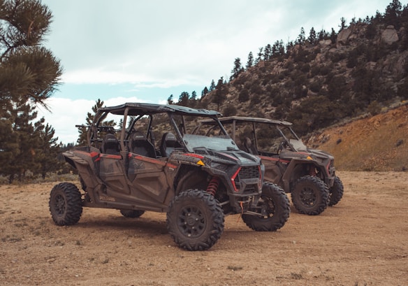 Two off-road vehicles are parked on a dirt path surrounded by a natural landscape featuring pine trees and rocky hills. The vehicles have rugged tires, open frames, and are designed for terrain exploration.