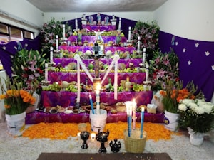 A colorful altar with several tiers is adorned with vibrant purple fabric, fruits, flowers, and candles. The arrangement includes various offerings like food, drinks, and decorative items, typical of a Day of the Dead ofrenda. Marigold petals are scattered on the ground, and tall white candles are placed throughout. A crucifix stands prominently in the center.
