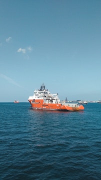 A large orange and white vessel navigates through calm blue waters, with additional ships visible in the distance against a clear sky.