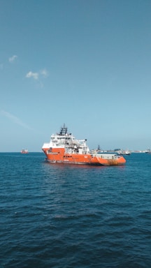 A large orange and white vessel navigates through calm blue waters, with additional ships visible in the distance against a clear sky.