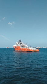 A large orange and white vessel navigates through calm blue waters, with additional ships visible in the distance against a clear sky.