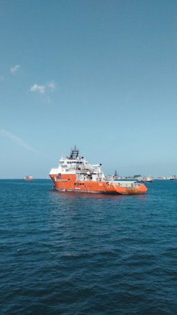 A large orange and white vessel navigates through calm blue waters, with additional ships visible in the distance against a clear sky.
