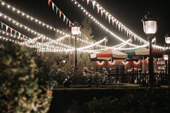 Strings of lights and festive bunting crisscross above an outdoor area, illuminating the scene. Vintage-style street lamps stand along the path, while tents with red, white, and green accents offer a classic, carnival-like atmosphere. Lush greenery and bushes frame the view.