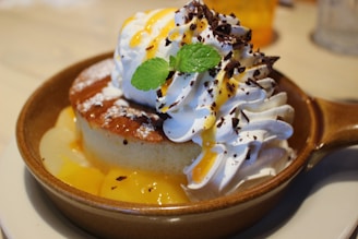 A delectable dessert featuring a golden-brown, fluffy pancake served with whipped cream, drizzled with caramel sauce, sprinkled with chocolate shavings, and garnished with a fresh mint leaf. The dish is presented in a rustic cream-colored bowl placed on a plate.