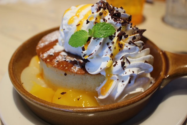 A delectable dessert featuring a golden-brown, fluffy pancake served with whipped cream, drizzled with caramel sauce, sprinkled with chocolate shavings, and garnished with a fresh mint leaf. The dish is presented in a rustic cream-colored bowl placed on a plate.