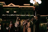 A group of musicians dressed in vintage-style clothing play brass instruments under streetlights. Behind them, a festive building is adorned with holiday lights and decorations.