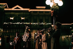 A group of musicians dressed in vintage-style clothing play brass instruments under streetlights. Behind them, a festive building is adorned with holiday lights and decorations.