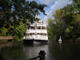 A large, white paddle steamer is navigating through a calm river surrounded by lush green trees. There are people on the deck enjoying the scenery. The sky is partly cloudy, adding a serene ambiance to the setting. A red and white buoy is floating nearby.