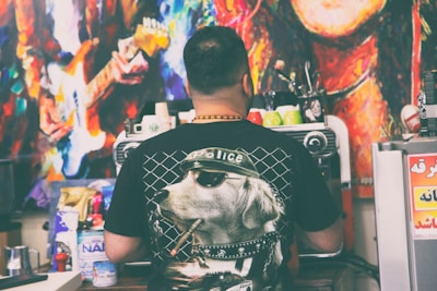 A person is standing with their back to the camera, wearing a black t-shirt featuring a graphic of a dog with sunglasses and a cap labeled 'Police'. They are in front of a coffee machine, with several colorful cups on top. The surrounding area includes various items and supplies, suggesting a café or coffee shop setting. In the background is a vibrant, abstract mural featuring a musician playing a guitar.