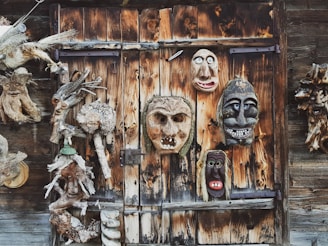 A wooden wall is adorned with an assortment of carved wooden masks and figures. The masks depict various facial expressions, ranging from grotesque to whimsical, and are mounted in an uneven pattern on the weathered wood backdrop. The textures and tones of the wood vary, showcasing natural knots, grains, and a range of browns and tans.