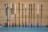 A row of floorball sticks, leaning vertically against a perforated wooden wall. The sticks have white handles and various colored blades, including pink, blue, and black shades. The floor is a smooth, greyish-blue surface.