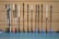 A row of floorball sticks, leaning vertically against a perforated wooden wall. The sticks have white handles and various colored blades, including pink, blue, and black shades. The floor is a smooth, greyish-blue surface.