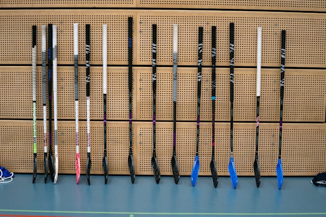A row of floorball sticks, leaning vertically against a perforated wooden wall. The sticks have white handles and various colored blades, including pink, blue, and black shades. The floor is a smooth, greyish-blue surface.