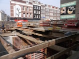 A construction site with a pit and wooden beams across the top. In the background, there are several multi-story brick buildings with a mix of white and dark facades. A billboard advertises a bakery with an image of a croissant.