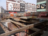 A construction site with a pit and wooden beams across the top. In the background, there are several multi-story brick buildings with a mix of white and dark facades. A billboard advertises a bakery with an image of a croissant.